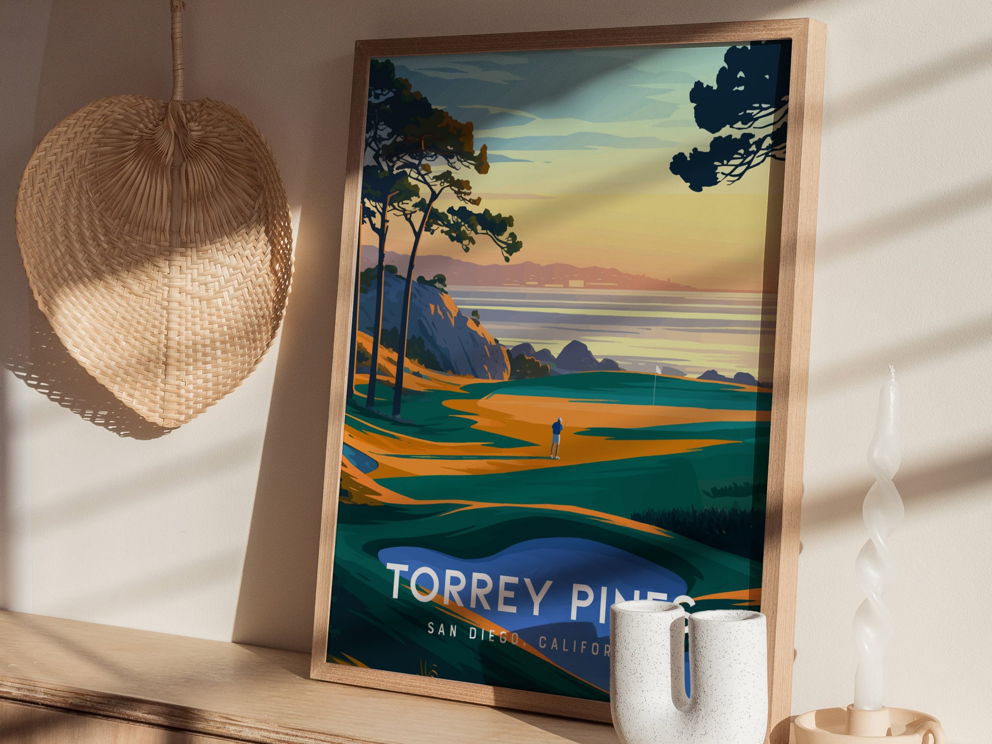 A framed poster of Torrey Pines Golf Course features vibrant coastal scenery with trees and cliffs, displayed on a wooden shelf beside a woven basket and decorative candle.