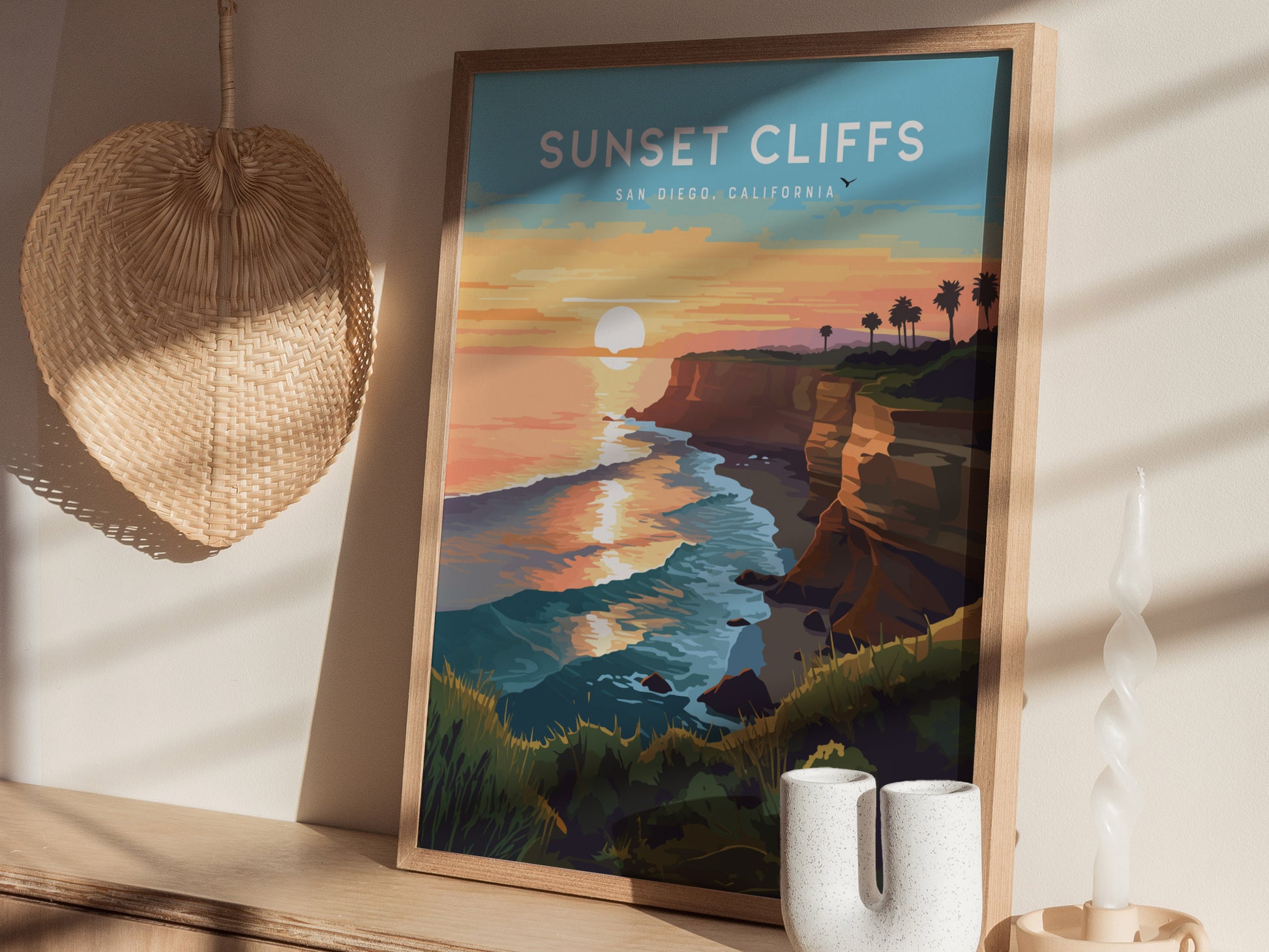 Framed artwork depicting Sunset Cliffs in San Diego with waves, cliffs, and palm trees under a sunset sky. The setting includes a woven wall decor and a unique candle holder.