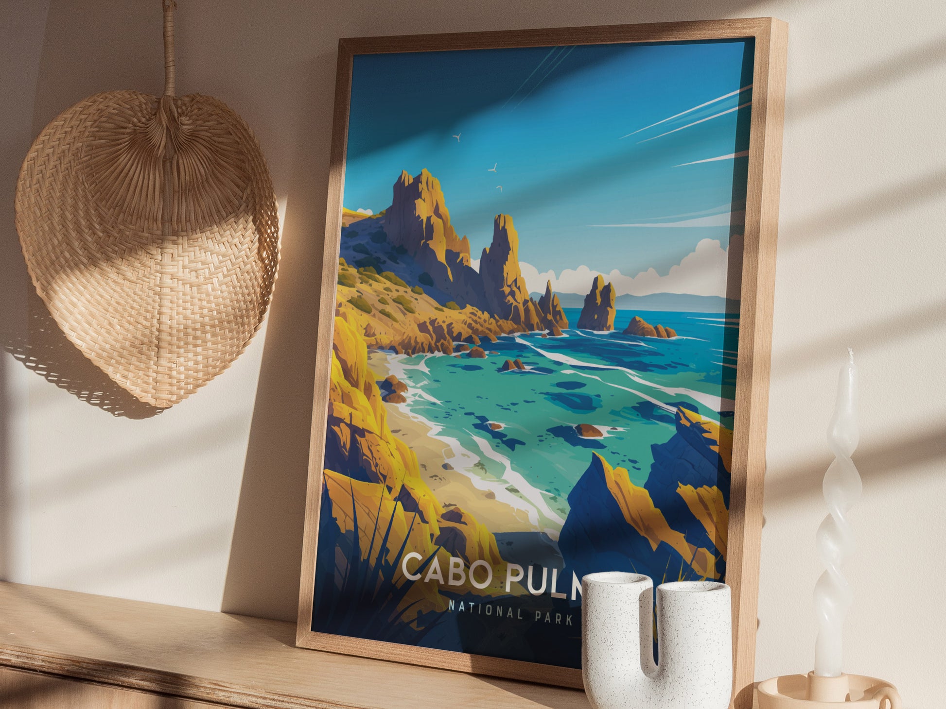 A framed poster depicting a vibrant coastal landscape with rocky cliffs and turquoise waters, titled Cabo Pulmo National Park, rests against a wall beside a woven basket and decorative candle.