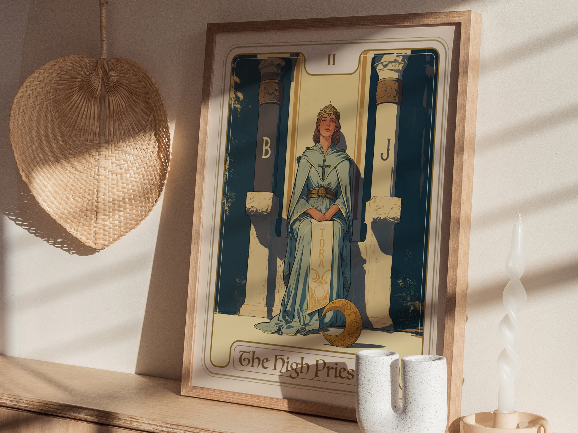 A framed High Priestess tarot card poster is displayed on a wooden shelf, featuring a serene figure between two pillars. Nearby are a woven fan, a twisted candle, and decorative.