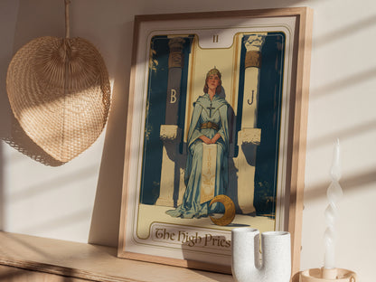 A framed High Priestess tarot card poster is displayed on a wooden shelf, featuring a serene figure between two pillars. Nearby are a woven fan, a twisted candle, and decorative.