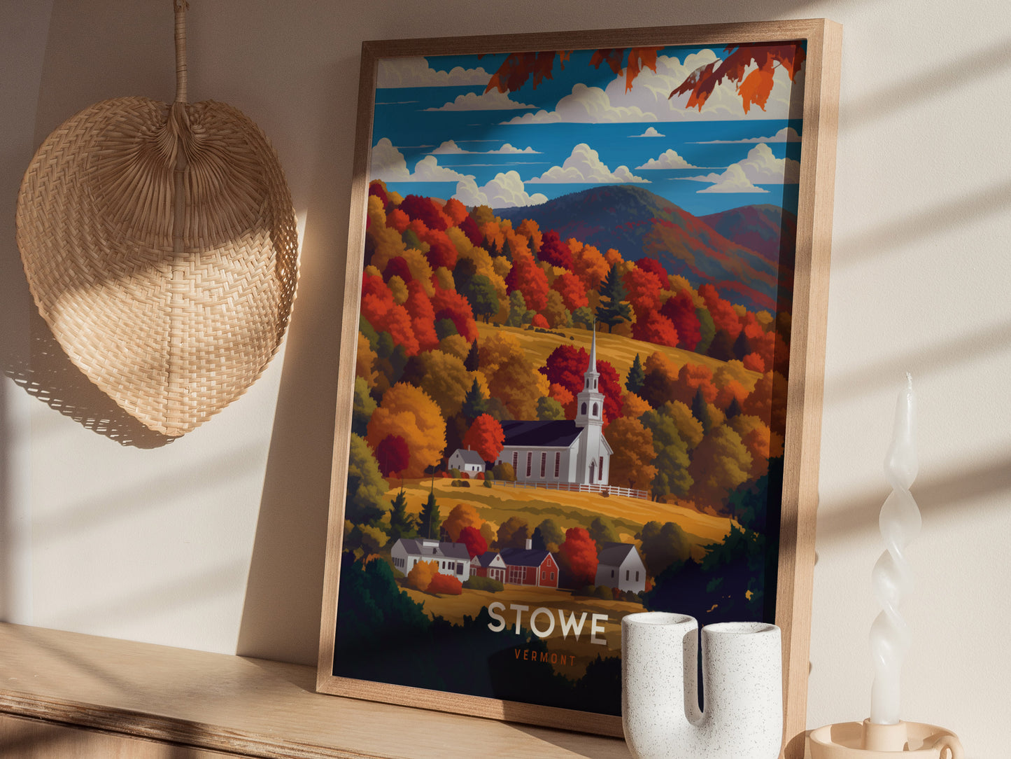 A framed poster of Stowe, Vermont, showcases a white church steeple amid vibrant autumn foliage and distant mountains. Its placed on a wooden shelf with a woven basket and decorative.