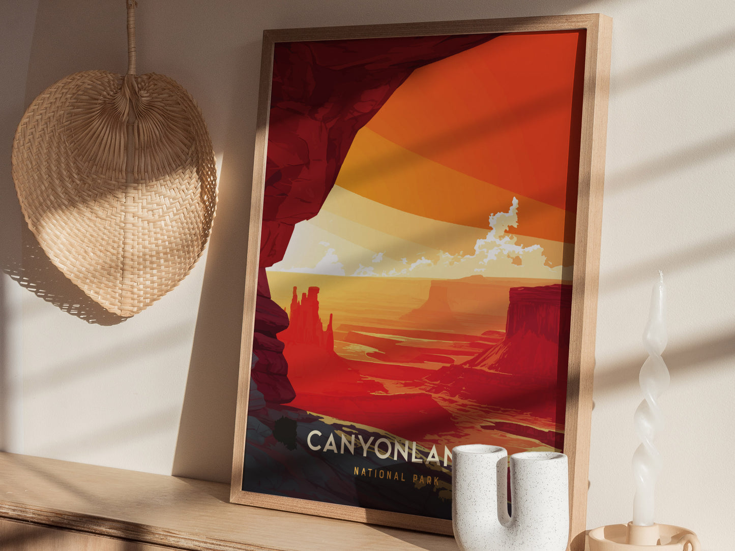 A framed Canyonlands National Park poster rests on a wooden shelf, accompanied by a woven fan and a spiral candle, casting soft shadows on the wall.