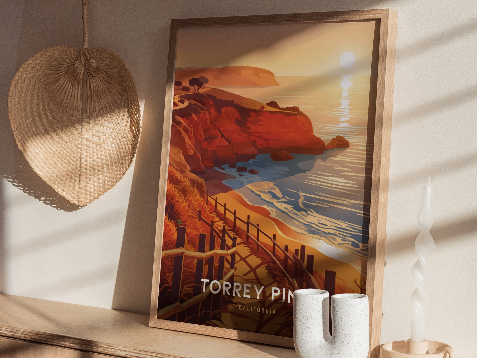 A framed poster depicting Torrey Pines coastal cliffs and ocean at sunset rests on a shelf. Nearby are a woven basket, spiral candle, and ceramic vase.