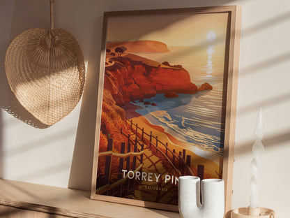A framed poster depicting Torrey Pines coastal cliffs and ocean at sunset rests on a shelf. Nearby are a woven basket, spiral candle, and ceramic vase.