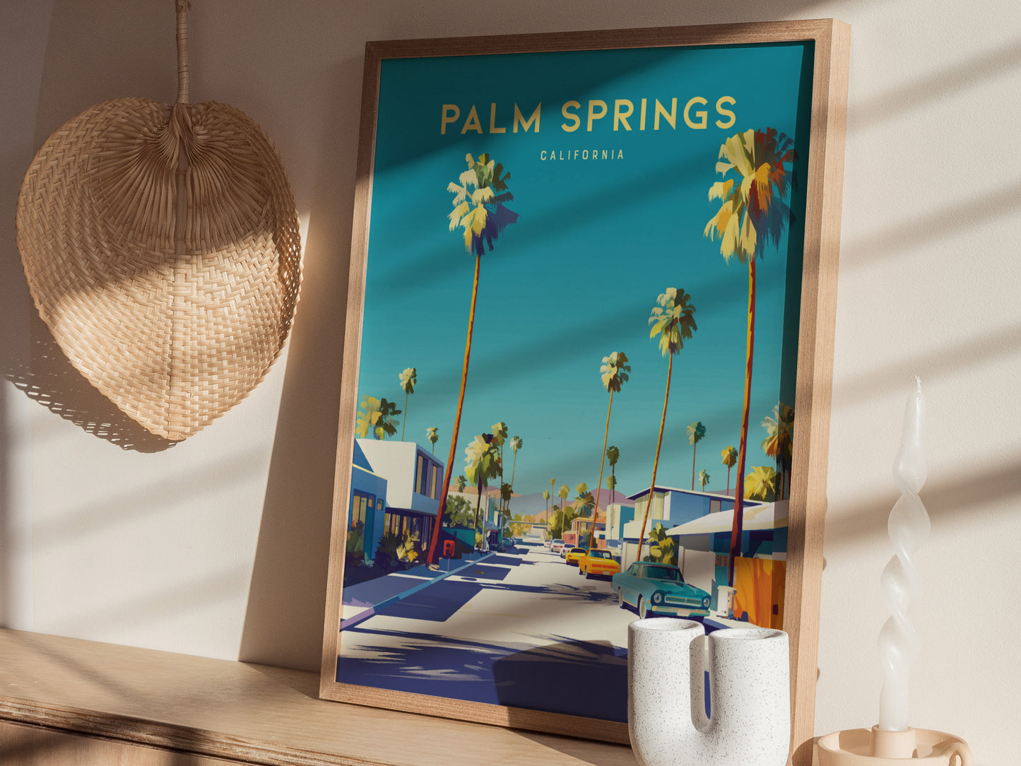 A framed poster of Palm Springs, California, featuring palm trees and mid-century architecture is displayed on a wooden shelf with a wicker basket and decorative candle nearby.