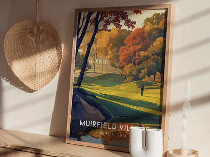 A framed poster of Muirfield Village Golf Club featuring vibrant autumn trees and a golfer, displayed on a wooden shelf with a woven basket and decorative candle nearby.