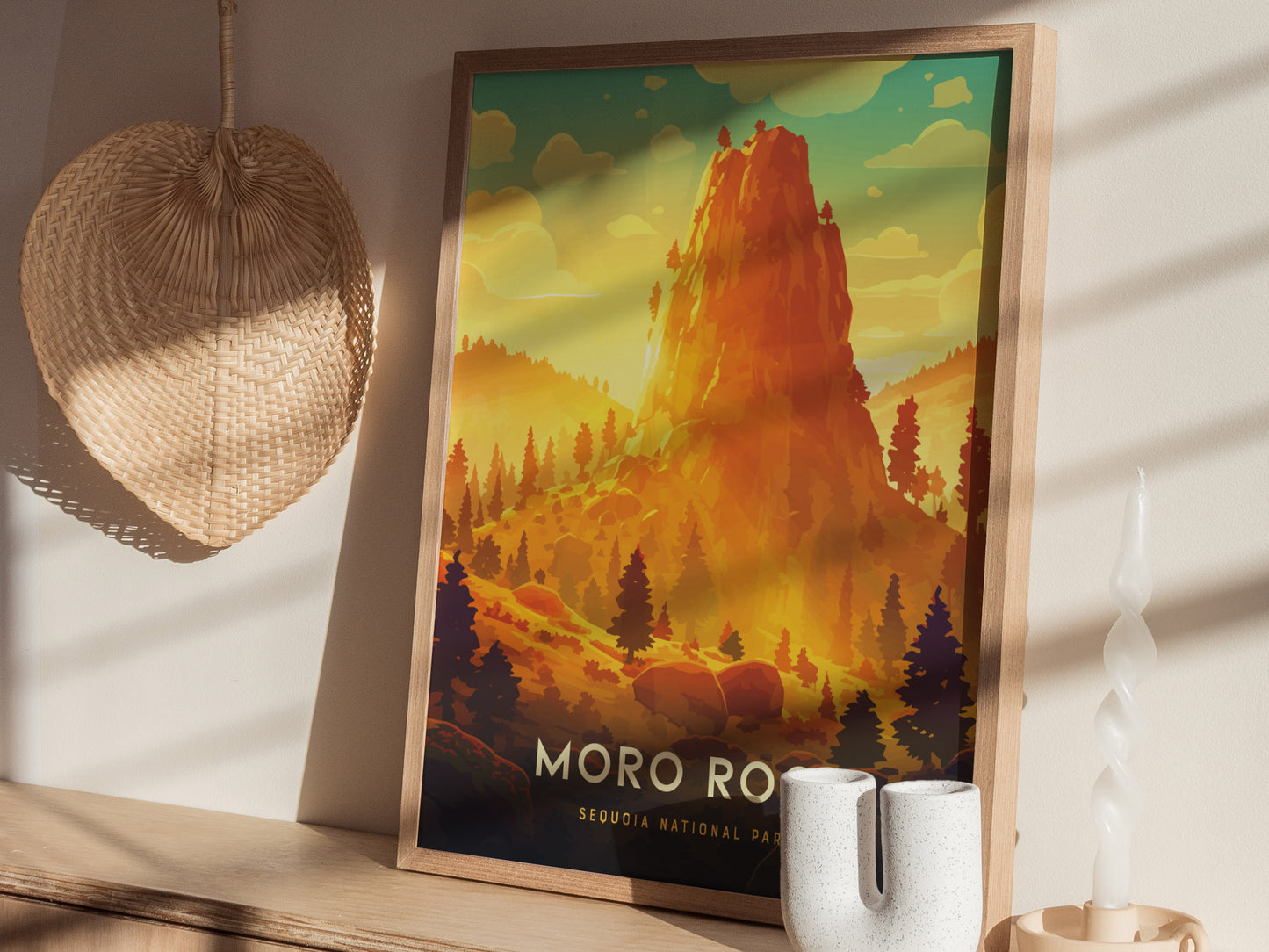 A framed Moro Rock poster depicting a vibrant, stylized landscape with a towering granite formation and forested hills. The image is set on a wooden shelf with decorative items nearby.