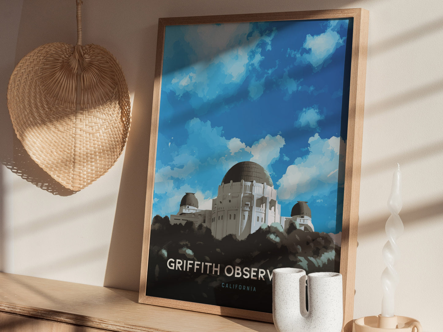 A framed Griffith Observatory poster sits on a wooden shelf, surrounded by a woven decorative piece and a uniquely shaped white candle, with sunlight casting shadows.