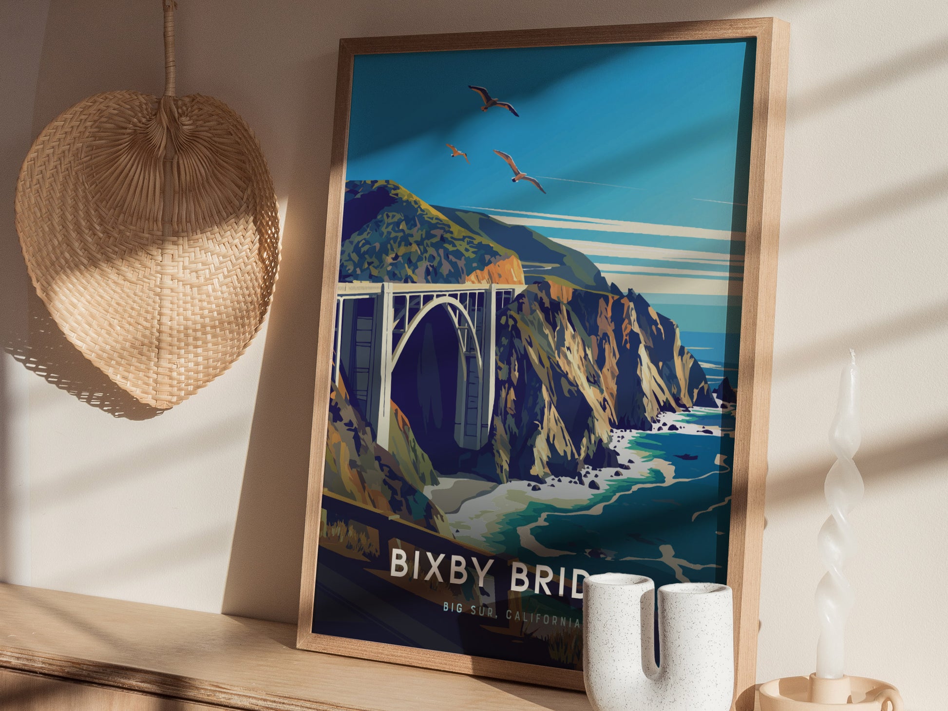 Framed poster of Bixby Bridge against a coastal landscape with seagulls flying. It sits on a wooden surface, accompanied by a woven basket and decorative white vases.