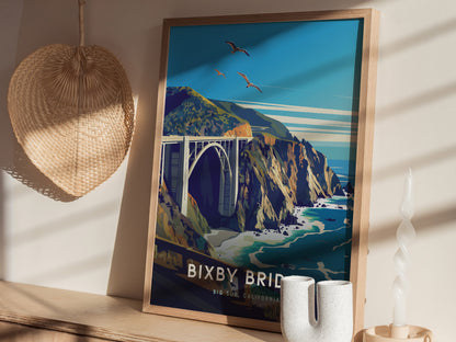 Framed poster of Bixby Bridge against a coastal landscape with seagulls flying. It sits on a wooden surface, accompanied by a woven basket and decorative white vases.