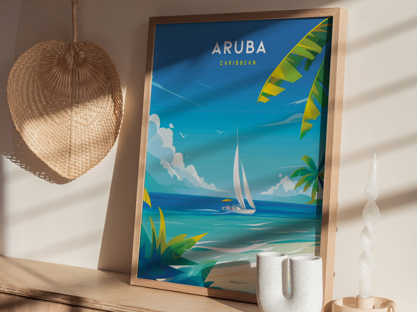 A framed Aruba travel poster featuring a sailboat on turquoise waters and palm leaves, displayed on a shelf with a woven wall hanging and decorative candle.