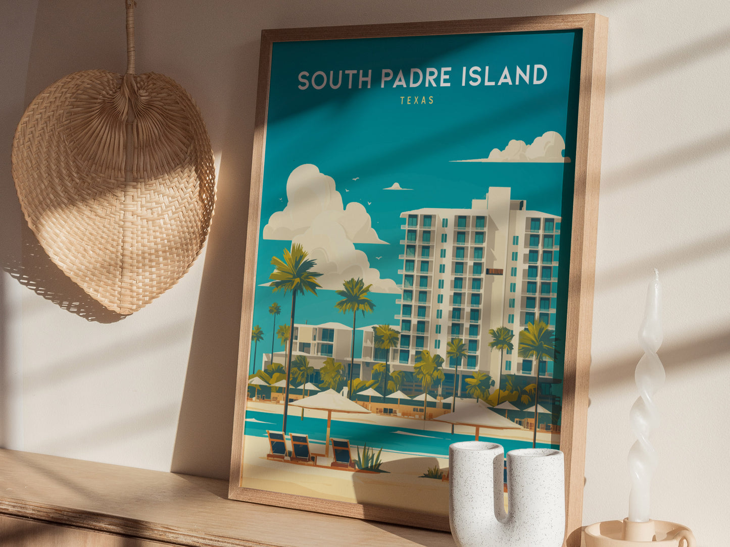 A framed South Padre Island poster depicting a coastal scene with palm trees and buildings is displayed on a wooden shelf, accompanied by a wicker fan and decorative candle.