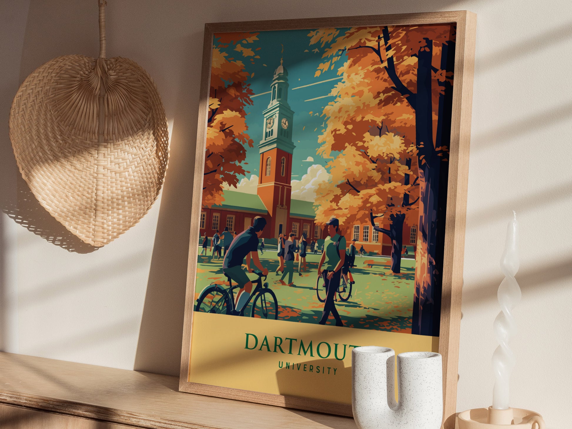 A framed Dartmouth University poster is displayed on a wooden shelf, surrounded by a wicker wall decoration and a white sculptural candle. The scene features students and autumn trees.