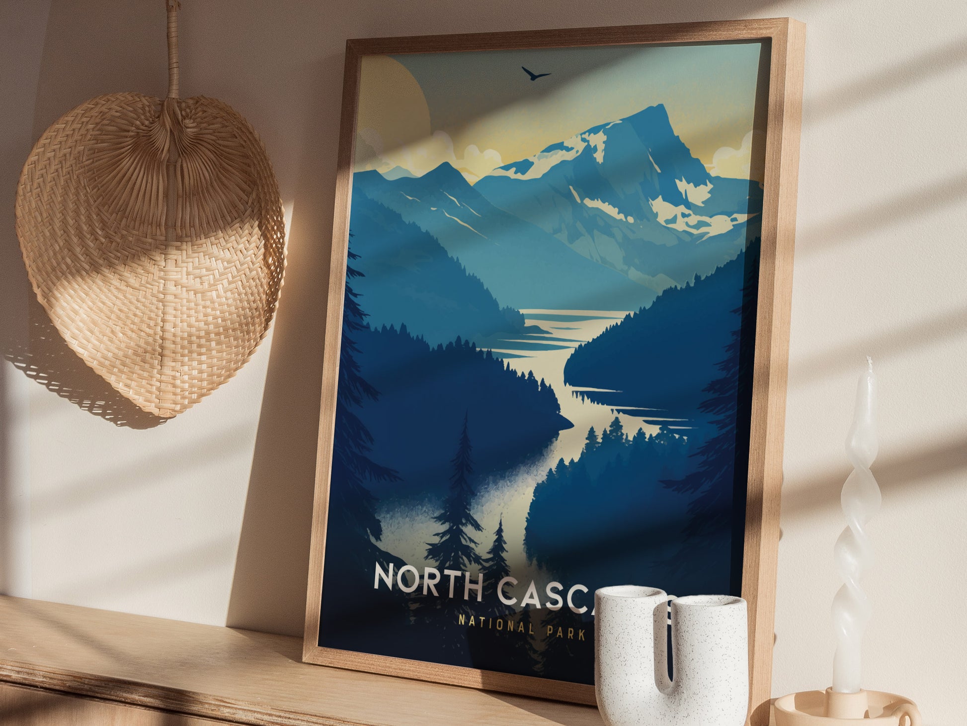 A framed poster of North Cascades National Park featuring blue mountains and a winding lake is displayed on a shelf, accompanied by a woven wall decor and a sculptural candle.