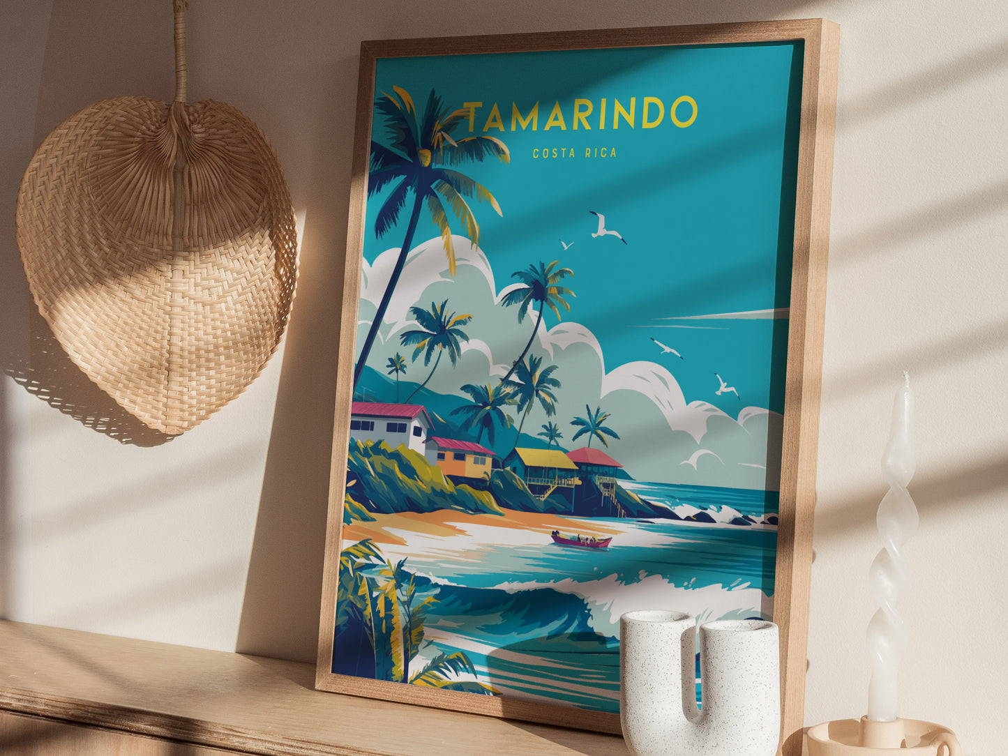 A vibrant poster depicting Tamarindo, Costa Rica, with palm trees, colorful houses, and seagulls over a beach scene is framed on a wooden surface. Nearby, a woven hat.
