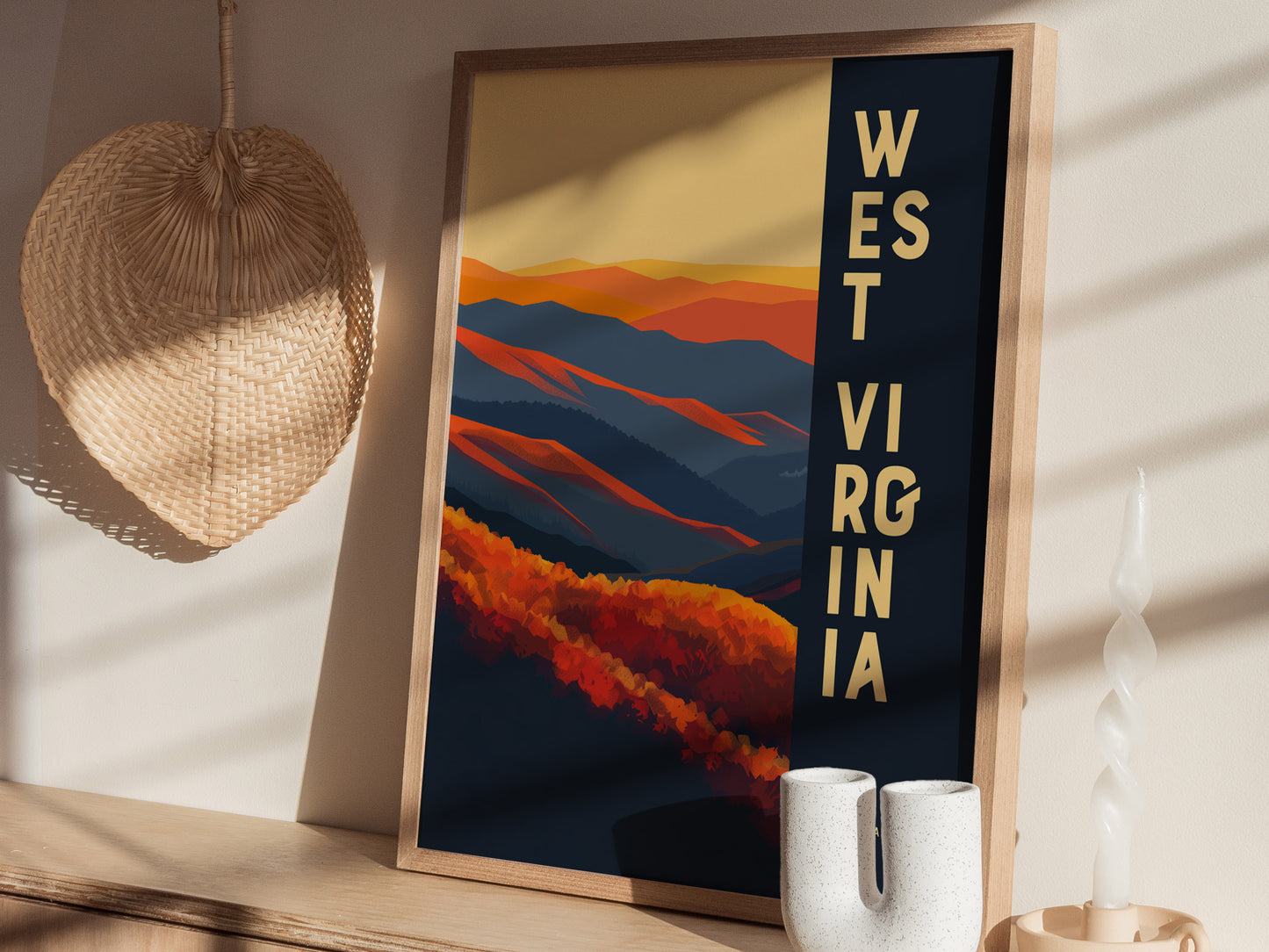 A framed West Virginia poster featuring abstract mountains in orange and blue tones is displayed on a wooden shelf beside a wicker decoration and a white twisted candle.