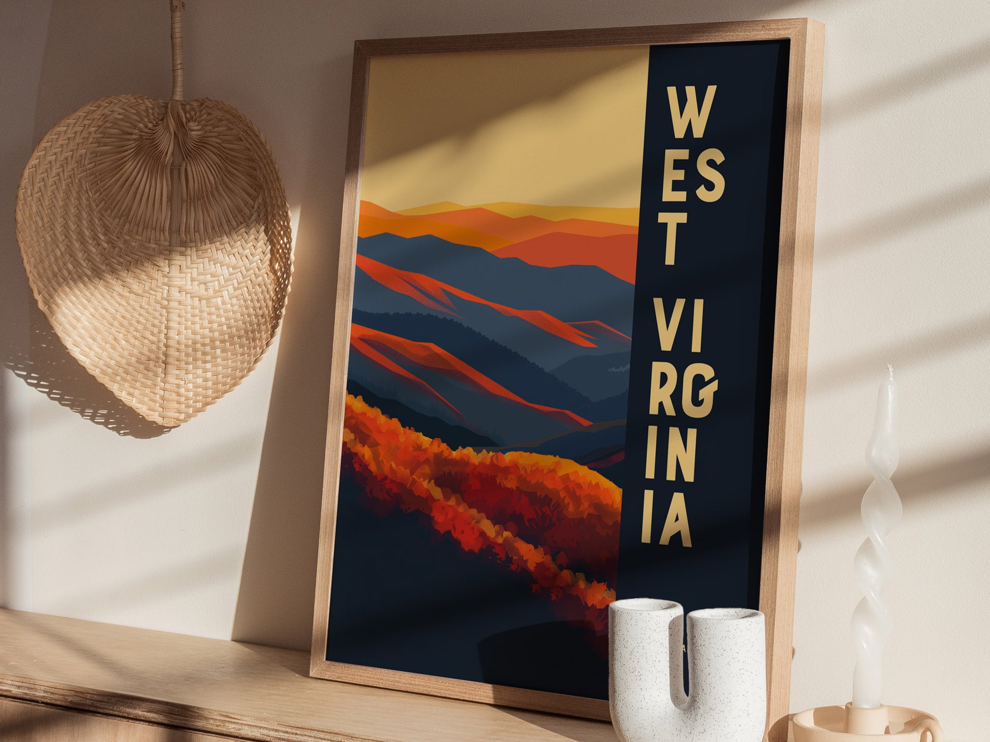 A framed West Virginia poster featuring abstract mountains in orange and blue tones is displayed on a wooden shelf beside a wicker decoration and a white twisted candle.