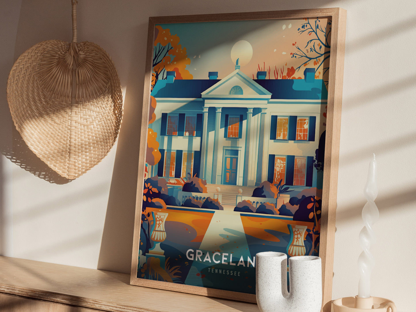 A framed Graceland poster depicting the iconic mansion is displayed on a wooden shelf beside a woven basket and decorative candles, bathed in warm sunlight.