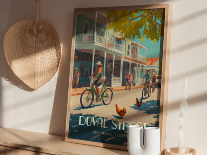 A colorful poster of Duval Street in Key West, featuring cyclists and roosters under shady trees, is displayed on a wooden shelf beside a wicker fan and decorative candle.