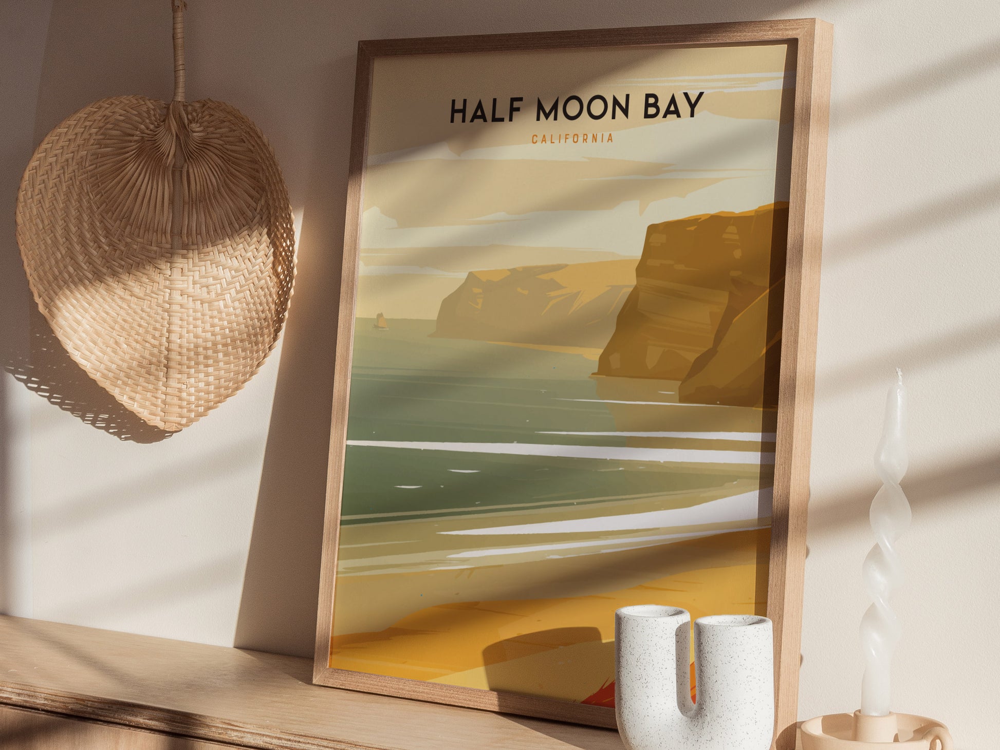 A framed Half Moon Bay, California poster with coastal artwork sits on a wooden shelf, accompanied by a woven basket and ceramic decor in a sunlit room.