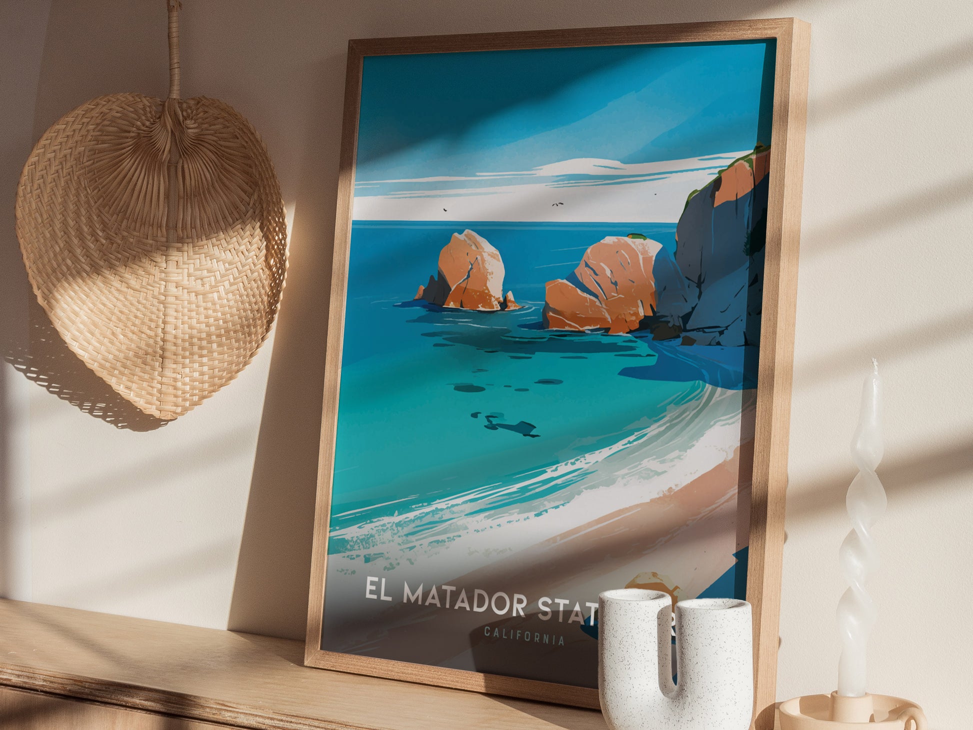 A framed poster of El Matador State Beach features vibrant blue ocean waves and rocky cliffs. Its displayed on a shelf beside a woven wall hanging and a white candle.