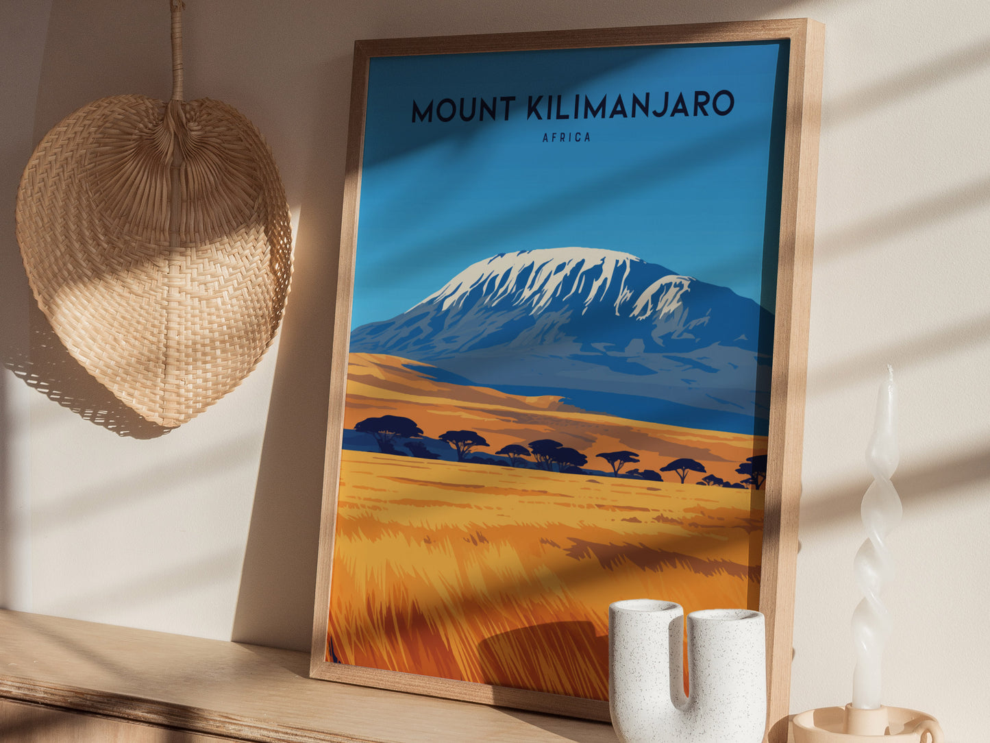 Framed poster of Mount Kilimanjaro with a snow-capped peak and savanna landscape, set against a wall with a woven basket and decorative candle.