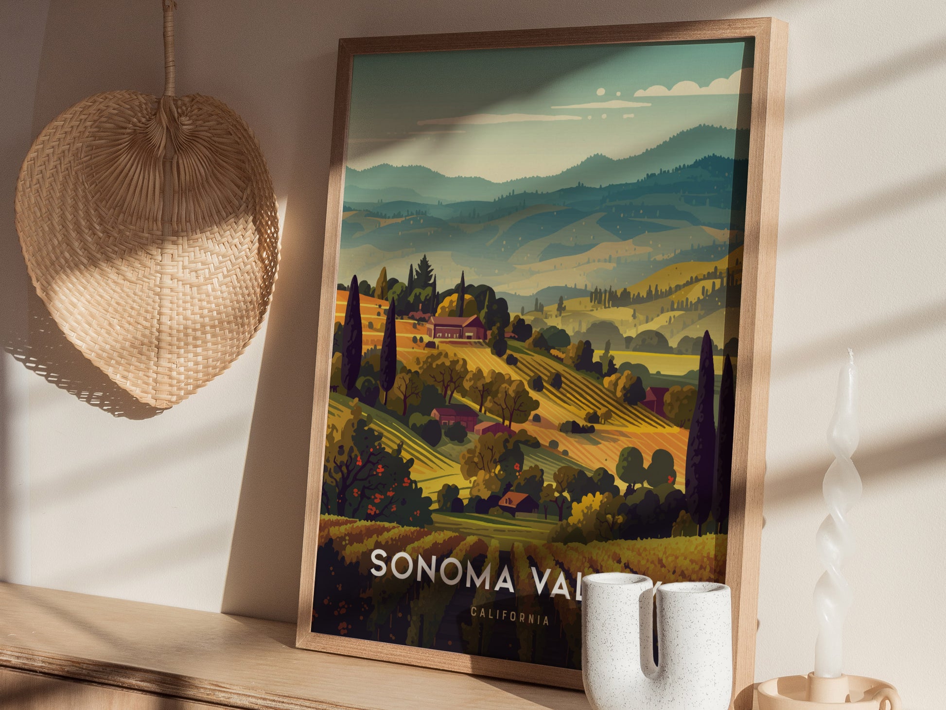 Framed Sonoma Valley poster depicting rolling vineyard hills with lush greenery and distant mountains, placed on a shelf next to a woven basket and a twisted white candle.