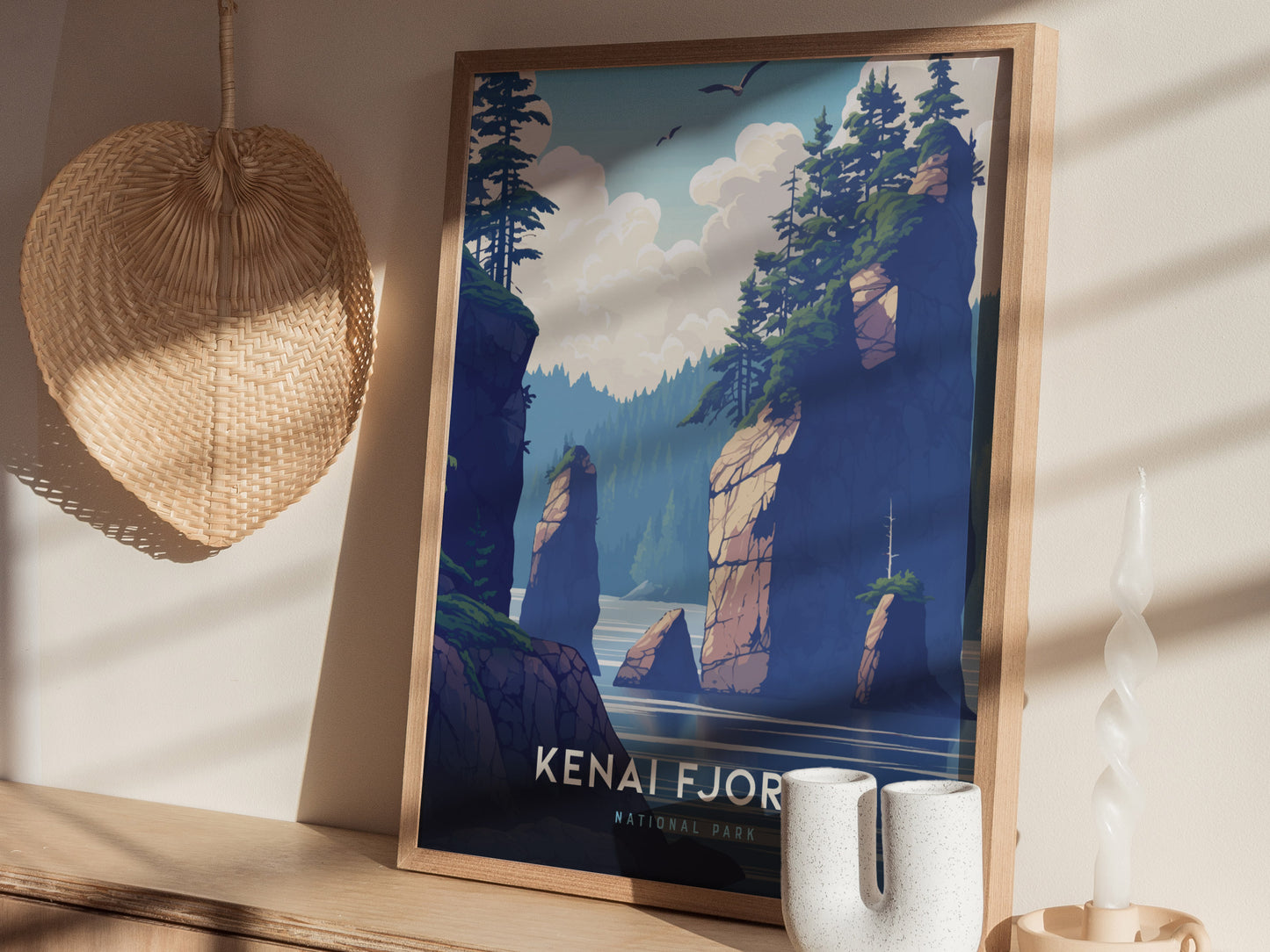 A framed Kenai Fjords National Park poster sits on a wooden shelf, depicting towering rock formations and lush greenery under a cloudy sky. A woven basket and candle are nearby.