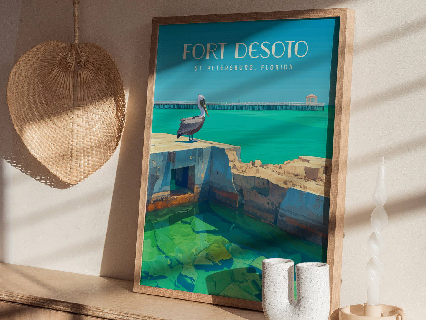Framed poster featuring a pelican on fort ruins with turquoise water, labeled Fort Desoto, St Petersburg, Florida, displayed on a wooden shelf beside a woven basket and twisted candle.