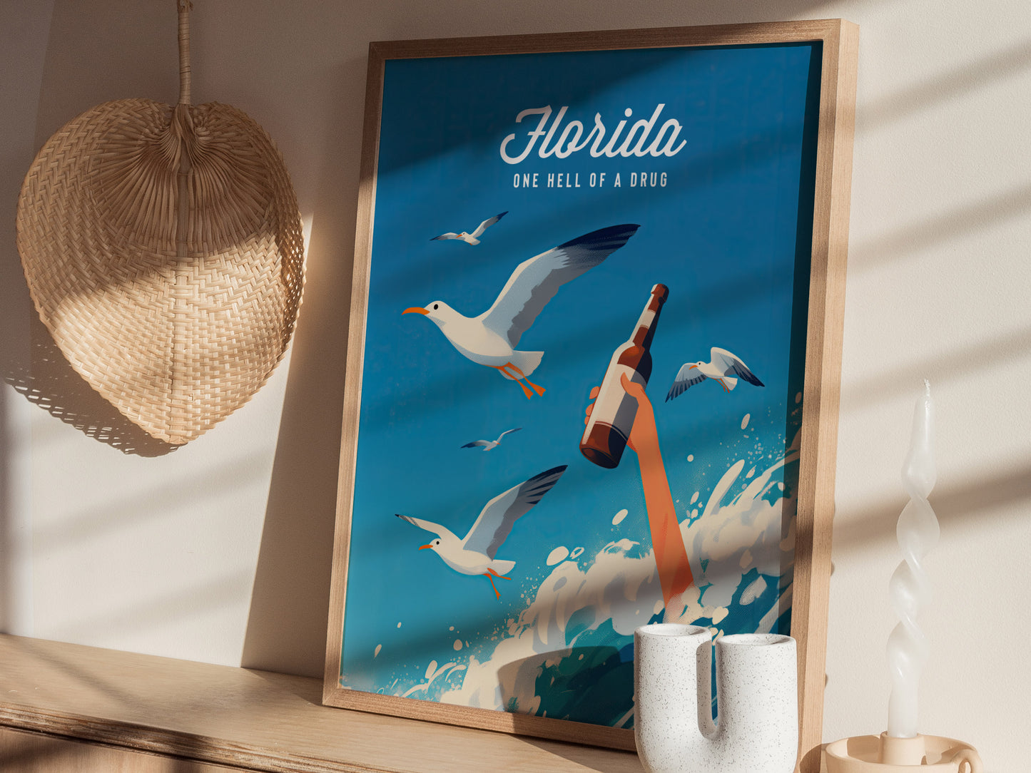 A framed poster titled Florida One Hell of a Drug features seagulls and a hand holding a bottle amid ocean waves. It leans against a wall near a decorative basket.