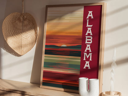 A framed abstract poster with ALABAMA text features warm sunset hues. Its placed on a wooden shelf with a wicker fan and a twisted candle nearby.
