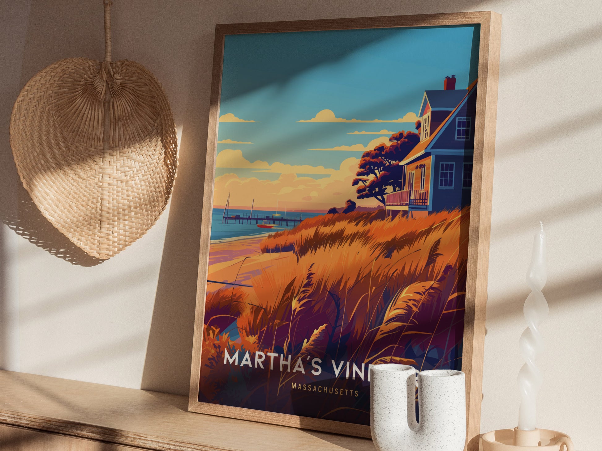 A framed poster of Marthas Vineyard is displayed on a wooden shelf, featuring a coastal scene with a house, grasses, and the ocean. A woven straw decoration hangs nearby.