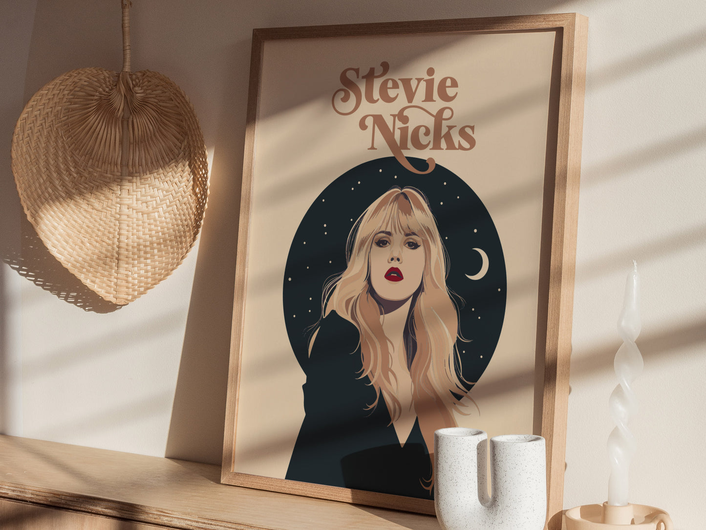 A framed poster of Stevie Nicks features an elegant portrait with celestial stars and a crescent moon. Its displayed on a wooden shelf alongside a woven fan and decorative items.