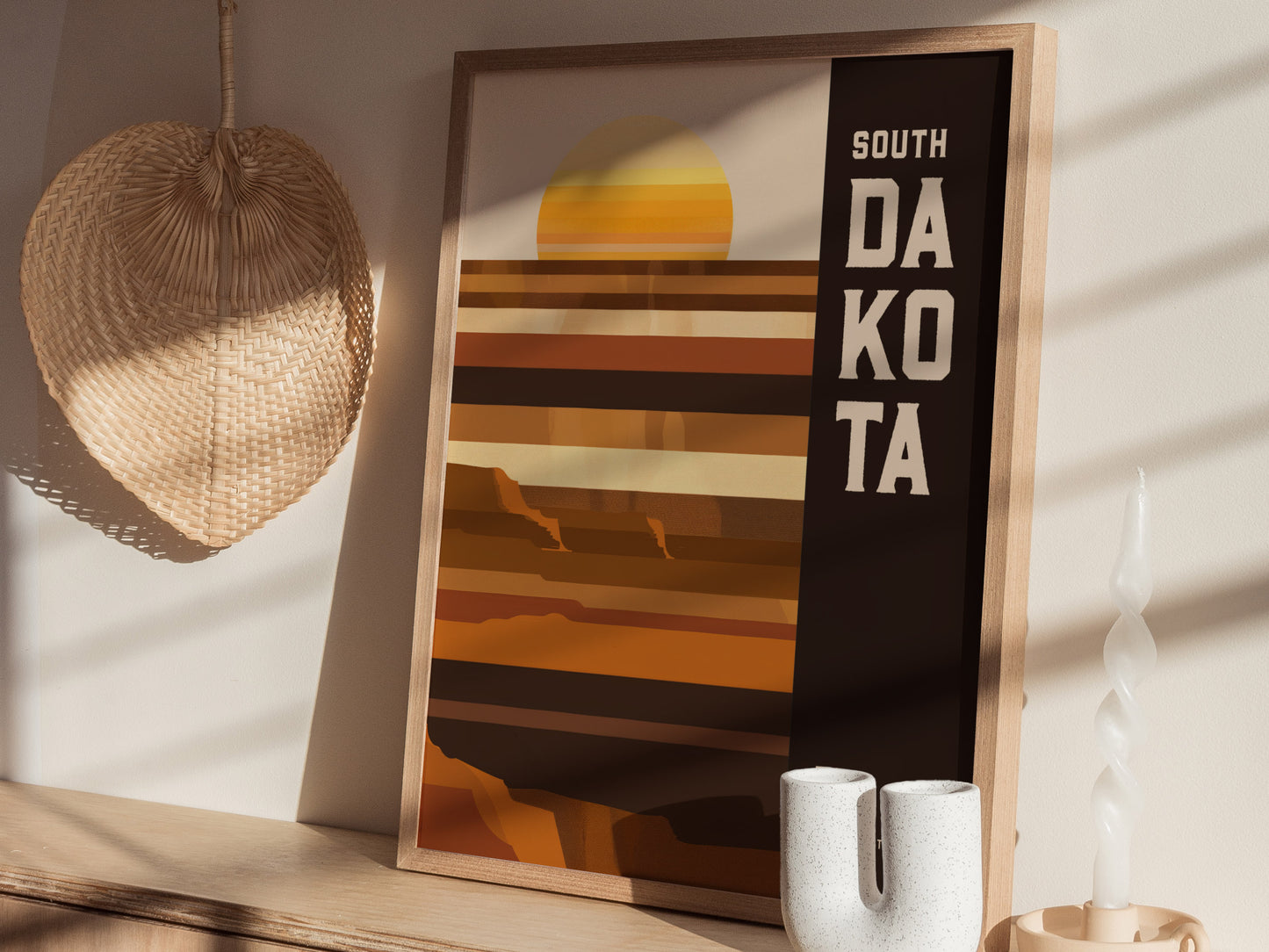 A framed South Dakota poster with a minimalist geometric design in earthy tones sits on a wooden shelf, next to a woven basket and a twisted candle.