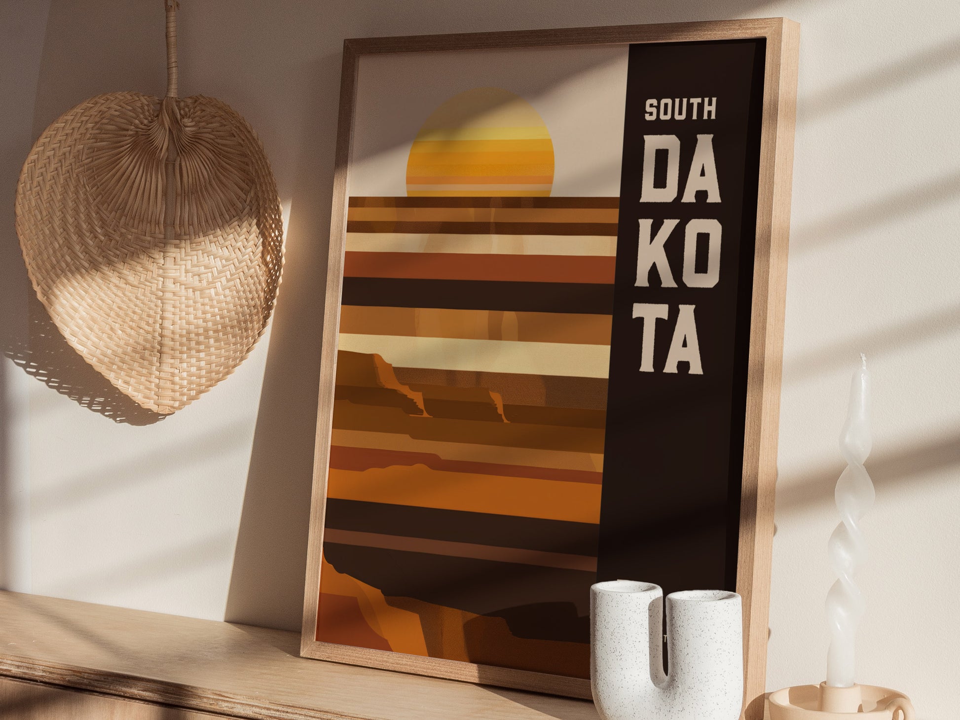 A framed South Dakota poster with a minimalist geometric design in earthy tones sits on a wooden shelf, next to a woven basket and a twisted candle.