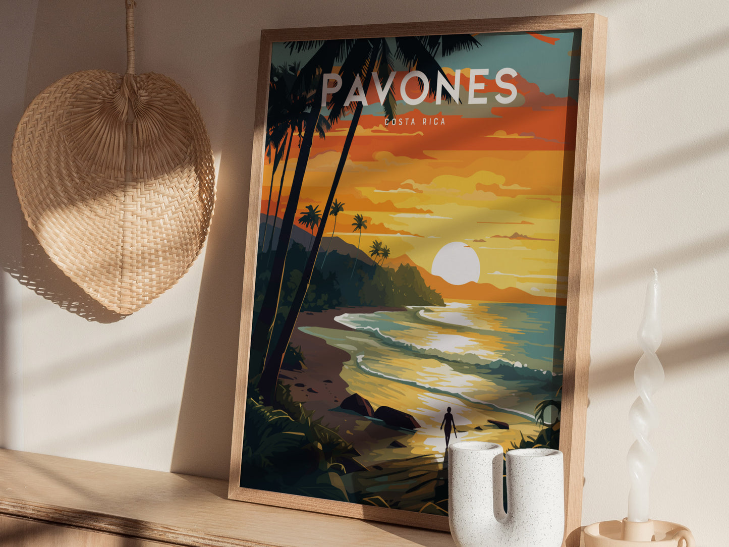 A framed surf poster depicting a sunset over a tropical beach with palm trees and waves. A straw hat and twisted candle add a coastal vibe to the setting.
