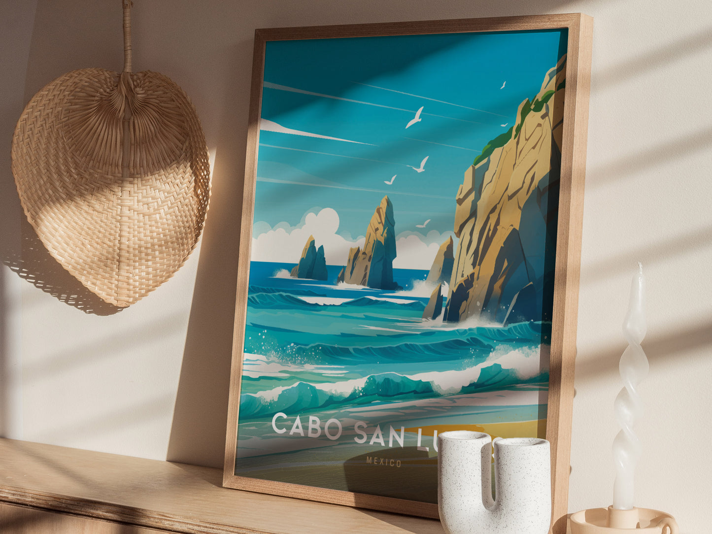 A framed poster of Cabo San Lucas featuring the Arch at Land’s End with turquoise waters and seagulls, set on a wooden shelf beside a woven basket and candle.