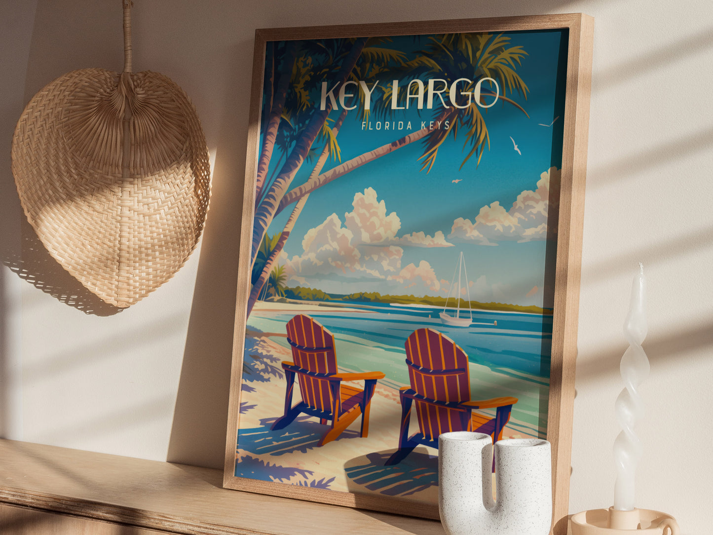 A framed Key Largo poster featuring two chairs on a beach under palm trees, with a sailboat on the horizon. The setup includes a woven fan and a twisted candle.