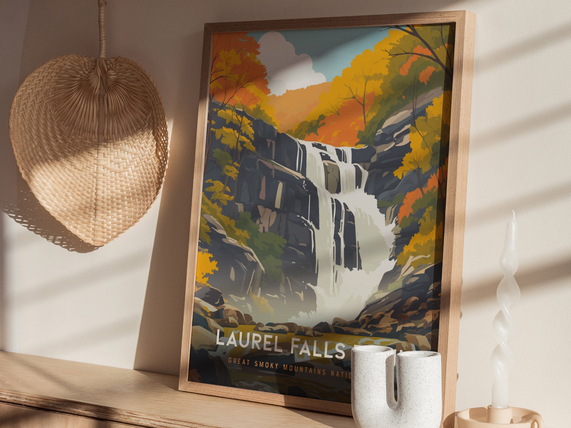 A framed poster of Laurel Falls, featuring a cascading waterfall surrounded by vibrant autumn foliage, is displayed on a shelf near a woven wall decoration and a decorative candle.