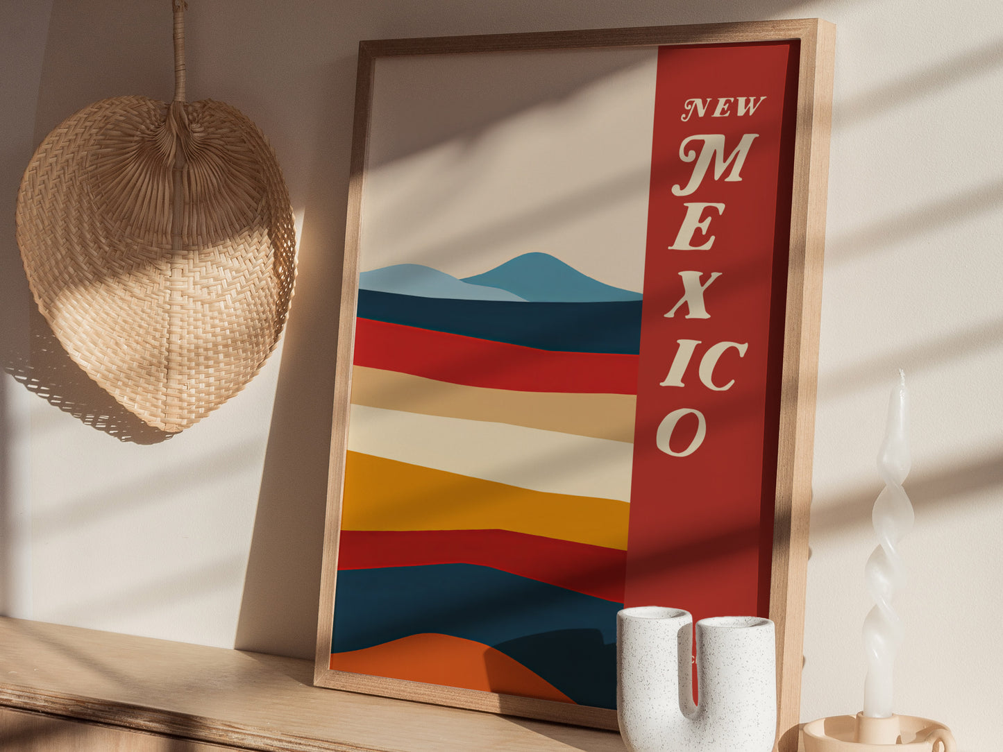 A framed poster with colorful abstract desert landscape and the text New Mexico is displayed on a wooden surface, accompanied by a woven fan and a twisted candle.