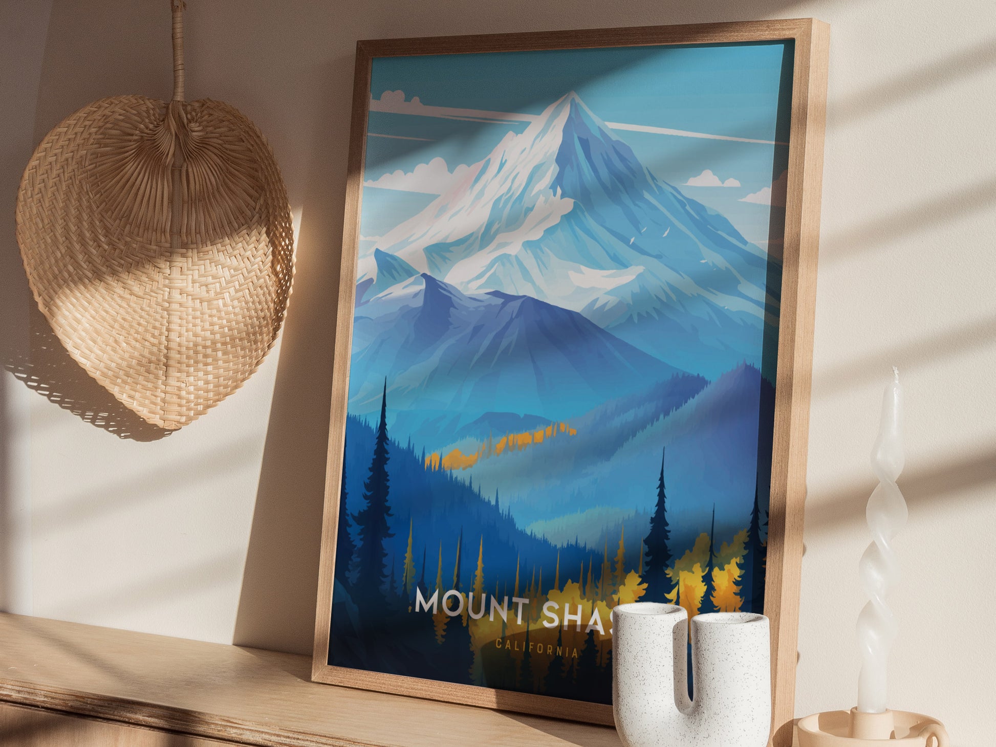 A framed Mount Shasta poster depicting a snow-capped mountain with blue and yellow forested foothills. It leans against a wall with a woven decoration and a twisted candle nearby.