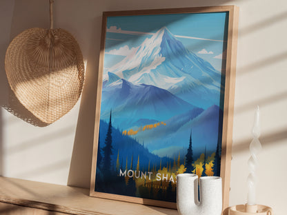 A framed Mount Shasta poster depicting a snow-capped mountain with blue and yellow forested foothills. It leans against a wall with a woven decoration and a twisted candle nearby.
