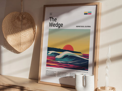 A framed poster titled The Wedge featuring a vibrant wave and sunset design sits on a wooden shelf alongside a woven fan and a twisted white candle.
