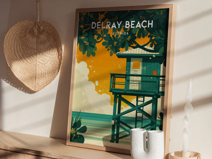 A framed Delray Beach poster with a vibrant lifeguard stand is displayed on a wooden surface, accompanied by a woven hat and decorative candle.