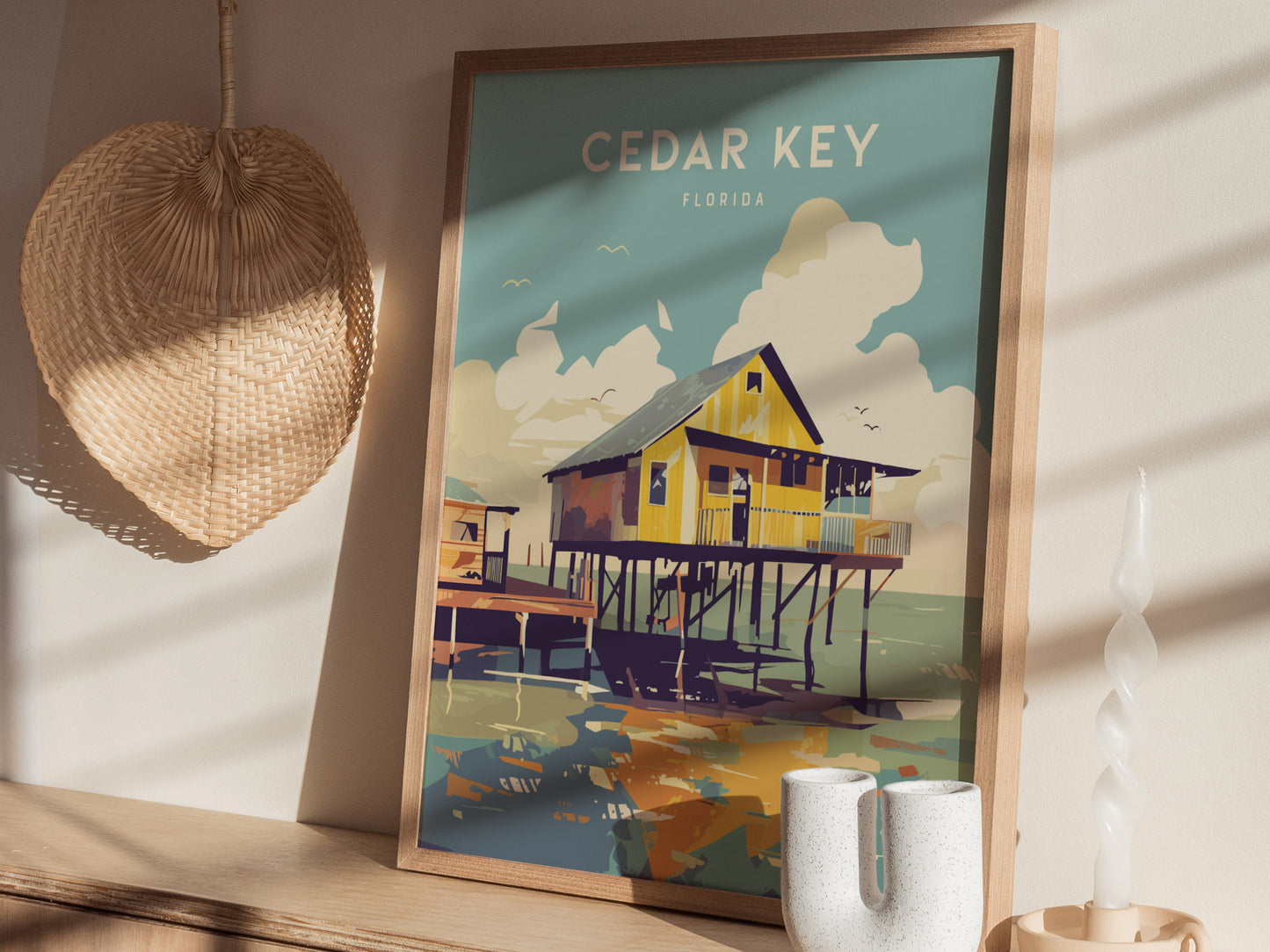 A framed Cedar Key, Florida poster features a stilt house over coastal waters, placed on a wooden shelf beside a woven basket and ceramic candle holder.