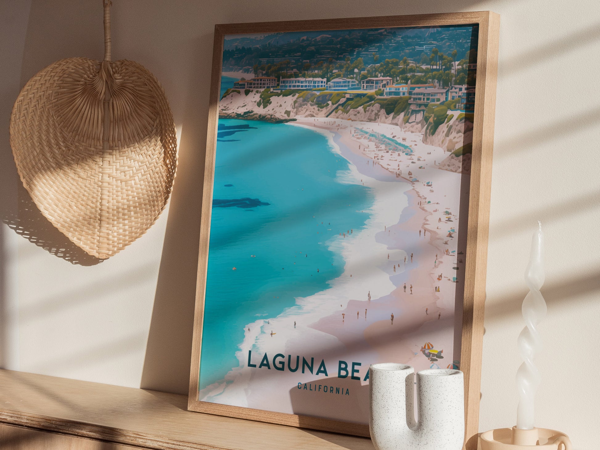 A framed poster of Laguna Beach, California, featuring a scenic coastal view with turquoise waters and sandy shores, is displayed on a wooden shelf beside a woven fan and candle.