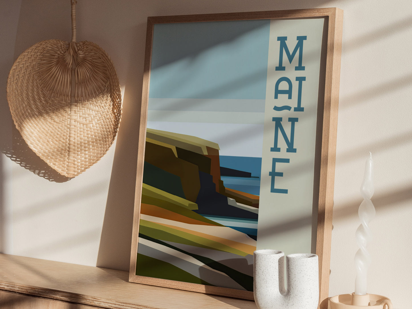 A framed Maine poster with abstract coastal scenery is displayed on a wooden surface. A woven basket and a twisted candle add decorative elements to the scene.