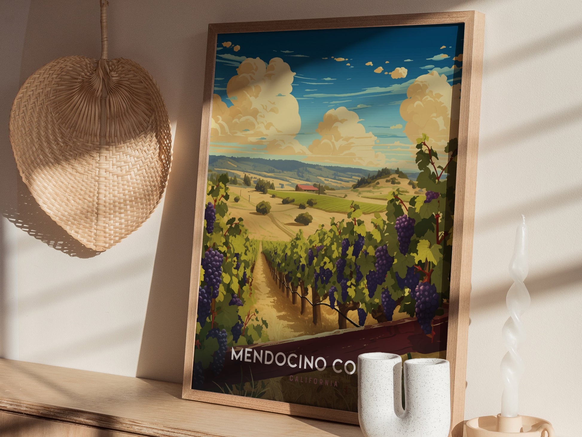 A framed poster of Mendocino County depicting a vineyard landscape with rolling hills under a blue sky. The setting includes a woven basket and decorative candle on a wooden surface.
