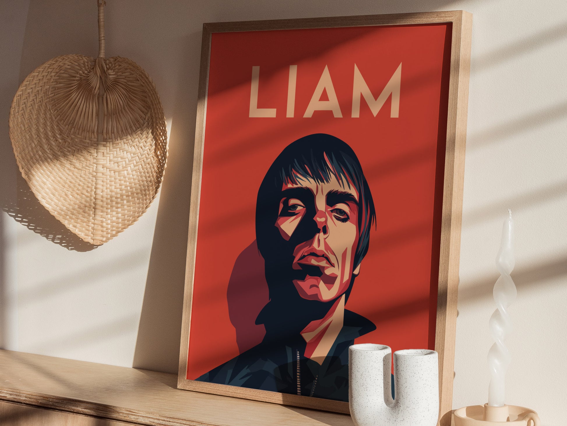 A framed poster with an artistic portrait and the name Liam on a red background sits on a wooden surface next to a woven fan and a twisted candle.
